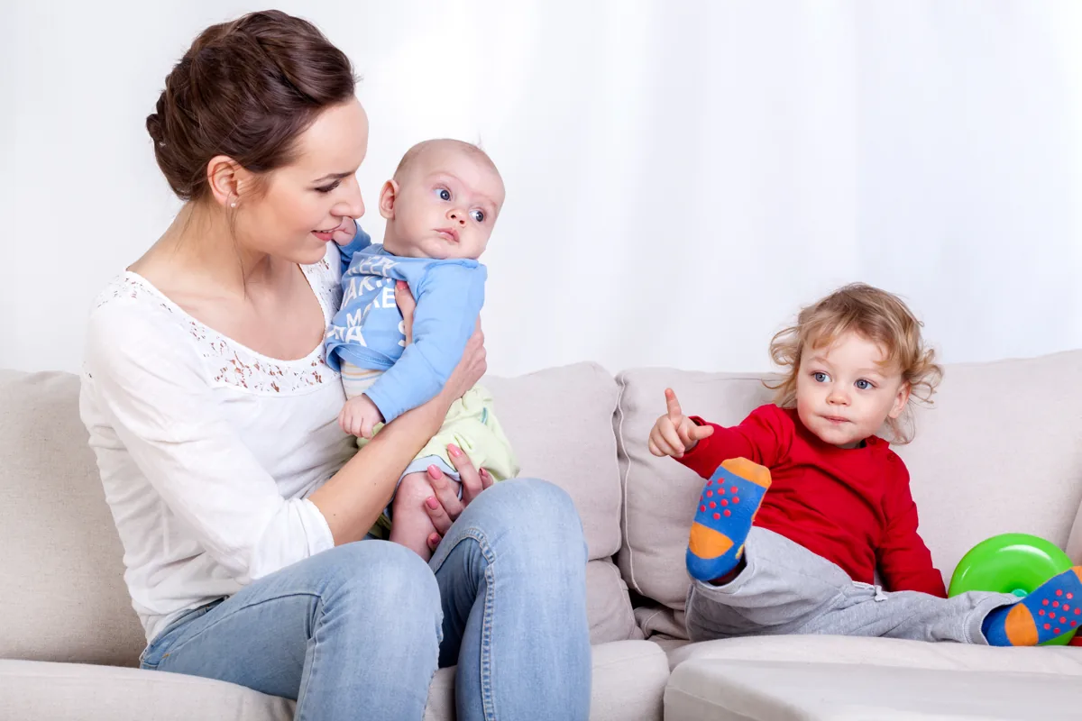 אמא יושבת על ספה ומחזיקה תינוק, ולצידה פעוט משחק בצעצוע